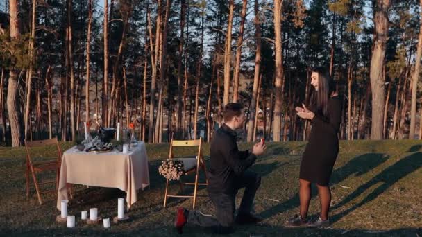 Allmän inställning med en ung man som friar i äktenskap sin älskade i naturen, mot bakgrund romantisk dekoration och granar. Den unga kvinnan säger ja — Stockvideo