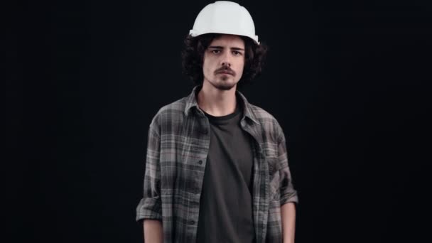 El joven trabajador con el dedo en los labios dice que se calle, en silencio. Lleva un casco de construcción y una camisa cerrada sobre un fondo negro en el estudio, concepto industrial, empleados — Vídeos de Stock