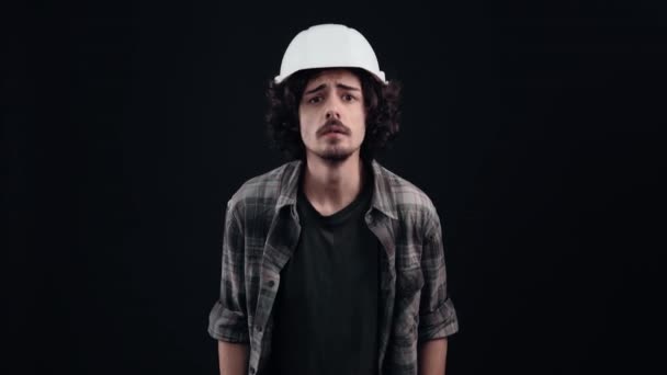 Young hipster shocked with his hands on his head says wow surprised. He wears a construction helmet and a closed shirt on a black background in the studio, Industrial concept, employees — ストック動画