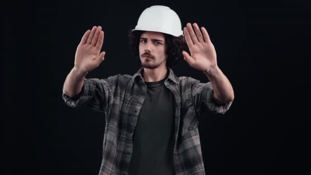 El joven guapo con las palmas emite una pared de cristal, mirando sus palmas. Lleva un casco de construcción y una camisa cerrada sobre un fondo negro en el estudio, concepto industrial, empleados — Vídeos de Stock