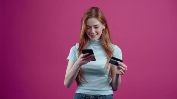 Szczęśliwa młoda kobieta z telefonem i kartą w ręku robi zakupy online, w niebieskiej, luźnej koszulce, odizolowanej na różowym tle. Koncepcja stylu życia narodów — Wideo stockowe