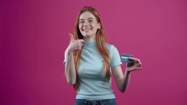 Happy redhead teenager with card in hand recommends online transactions, in blue casual t-shirt, isolated on pink background. The concept of peoples lifestyle — Stock Video