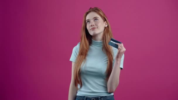A vörös hajú fiatal nő, aki a kártyával a kezében gondolkodik, a következő vásárláson gondolkodik, egy kék alkalmi pólóban, rózsaszín alapon elszigetelve. A népek életmódjának fogalma — Stock videók
