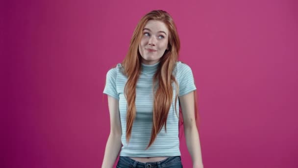 Η φακιδοκοκκινομάλλα τρέμει αβέβαια για την απάντησή της και χαμογελά πλατιά, αλλάζοντας το βλέμμα της προς διαφορετικές κατευθύνσεις, με ένα μπλε casual T-shirt, απομονωμένη σε ροζ φόντο. Η έννοια των λαών — Αρχείο Βίντεο