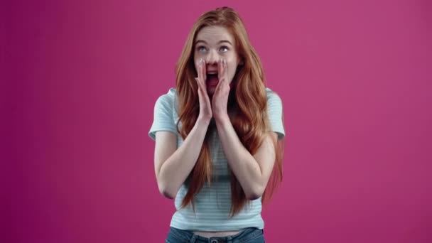 The red-haired and freckled teenager whispers a secret isolated from foreign ears by placing her hands on her mouth and then smiles innocently, in a blue casual T-shirt, isolated on a pink background — Stock Video