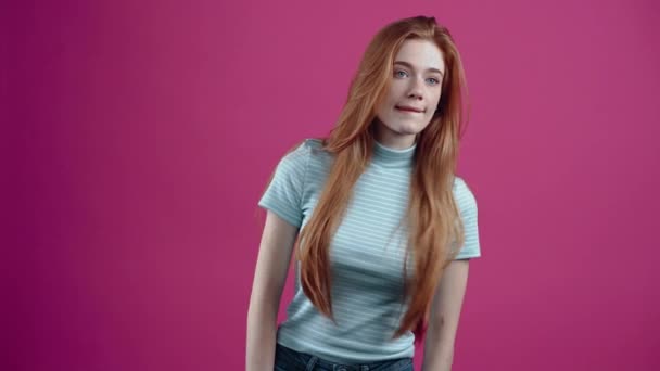 The concentrated young redhead looks for the right person, and after finding her, she greets her with his hand, and smiles at her, in a blue casual T-shirt, isolated on a pink background. The concept — Stock Video