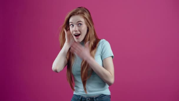 赤い髪のティーンエイジャーは、ピンクの背景に隔離されたカジュアルな青のTシャツで、彼女を驚かせ、彼女の笑顔を作る新しい噂を聞きます。人々の生活様式の概念 — ストック動画
