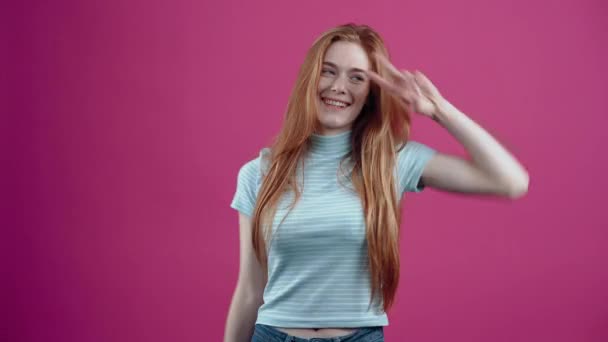 De roodharige party girl beweegt zich naar het ritme van de muziek, improviseren dans moves, in een blauw casual T-shirt, geïsoleerd op een roze achtergrond. Het concept van de levensstijl van mensen — Stockvideo