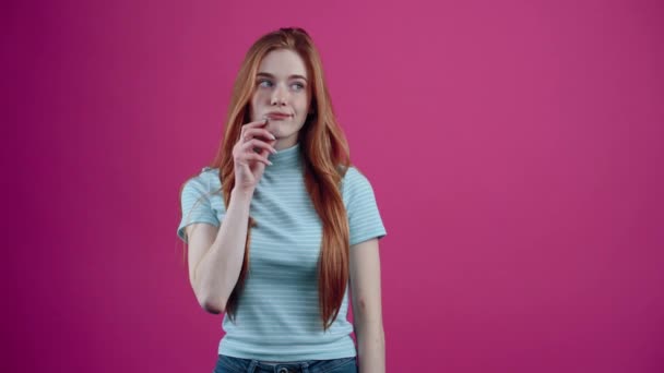 Rudovlasá, zamyšlená dívka přemýšlí o nápadu, s rukama přejíždějícíma kolem úst, v modrém neformálním tričku, izolovaném na růžovém pozadí. Koncept životního stylu lidí — Stock video