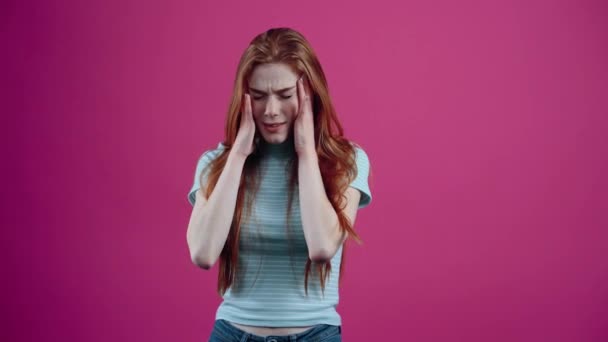 強調された赤い髪の若い女性は、疲労感の兆候として彼女の頭に彼女の手を置き、ピンクの背景に隔離された部分に失望しているように見えます。人々の生活様式の概念 — ストック動画