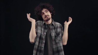 Charismatic, dreamy young man, shrugs and raises his hands in ignorance, then points his index finger to his chin, looking to one side, looking for a solution. Isolated on black background, Concept of