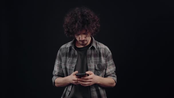 The charismatic young man, seriously, types angrily on the phone, after which he surfs the internet. Isolated on black background, Concept of life. Peoples emotions. 4k portrait — Αρχείο Βίντεο