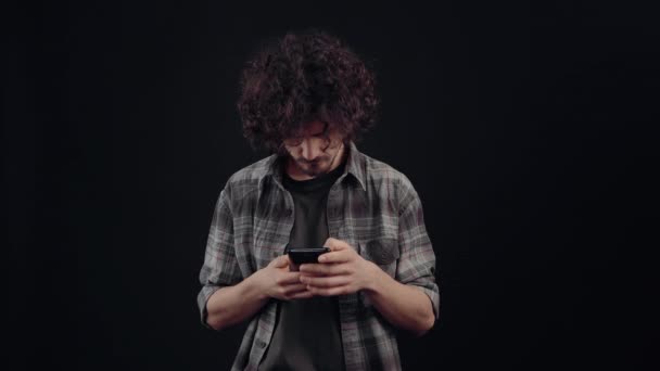 Харизматичный молодой человек, серьезно, печатает сообщение по телефону. изолированы на черном фоне, концепция жизни. эмоции людей. Портрет 4k — стоковое видео