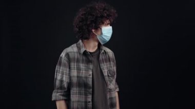 The young man with curly hair wears a mask, looks to one side and waves his hands in a sign that the others keep their distance from him. Isolated on black background, Concept of life. Peoples