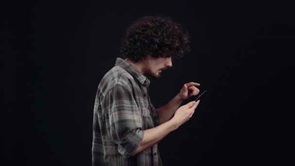 The optimistic young man, looking in profile on the phone screen, gets good news that makes him very happy, shouts wow and makes him show the like sign. Isolated on black background, Concept of life — Vídeo de stock
