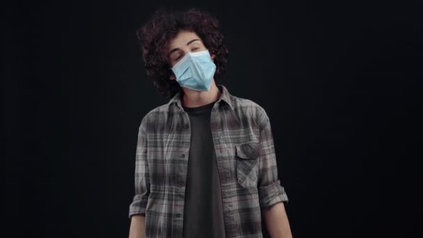 The young man with curly hair wears a mask, opens his eyes in surprise and then walks them in parts and then closes them in agreement. Isolated on black background, Concept of life. Peoples emotions — ストック動画