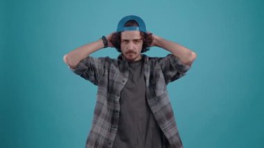 The charismatic young man, puts cap on his head, points forward saying, you, yes, you, arranges the hair and then confidently crosses his hands to chest, raising his eyebrows. isolated over turquoise