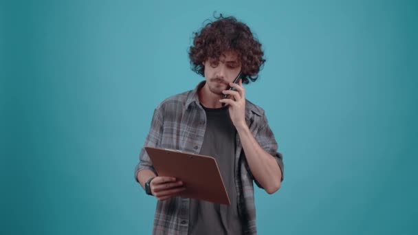 The charismatic and serious young man talking on the phone looks over the notes if he wrote down the information correctly. Isolated on a turquoise background. Concept of life. Peoples emotions. 4k — Αρχείο Βίντεο