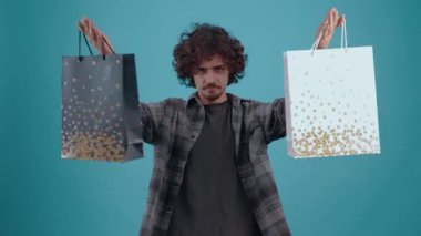The happy young man, after shopping, picks up two packages, looks at one and the other, nods approvingly. Isolated on a turquoise background. Concept of life. Peoples emotions. 4k portrait