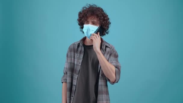 The boy with curly, charismatic hair talks on the phone with the mask on his face and moves in a good mood. Isolated on a turquoise background. Concept of life. Peoples emotions. 4k portrait — Wideo stockowe