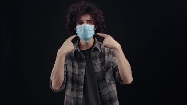 Młody człowiek z kręconymi włosami nosi maskę, wskazuje palcami wskazującymi na maskę i tym podobnym znakiem pokazuje potrzebę i troskę, aby ją nosić, na końcu wkłada palec do policzka — Wideo stockowe