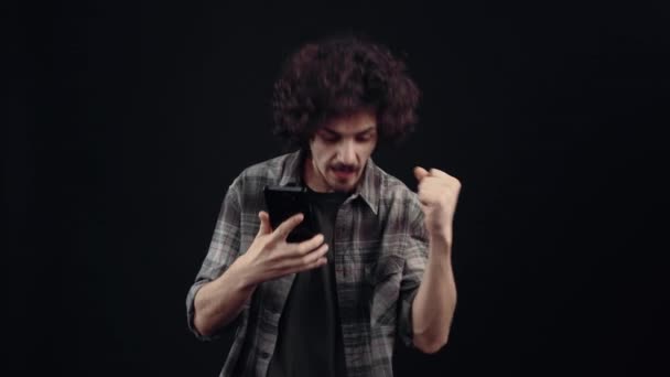 The charismatic, focused young man looks into the phone, receives news that makes him raise his arms victoriously and show the like sign. Isolated on black background, Concept of life. Peoples — Stock Video