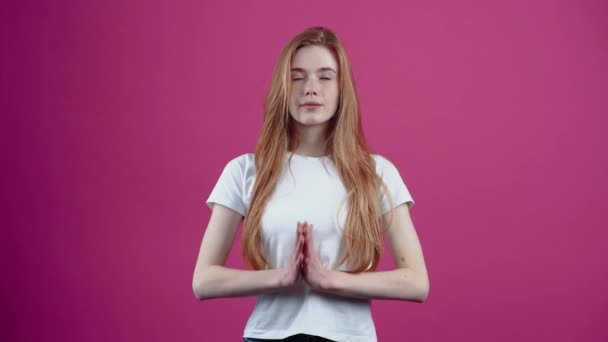 Portret van een mooie jonge vrouw die mediteert met haar handen tegen haar borst. Sproeten tienermeisje in een wit T-shirt, geïsoleerd op een roze achtergrond. Het concept van de levensstijl van mensen — Stockvideo