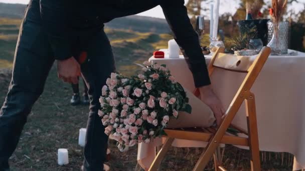 Den unga förälskade mannen erbjuder sin flickvän en stor bukett blommor, isolerade i naturen vid en romantisk middag mitt i solnedgången — Stockvideo