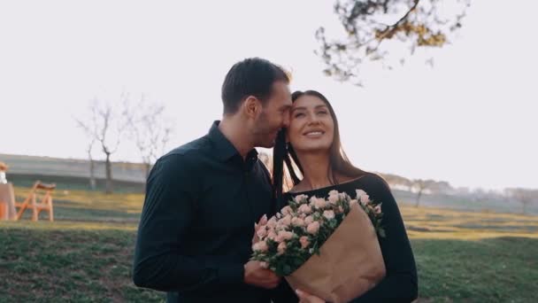 The happy couple, isolated in a dream meadow, at sunset, sit together and smile in love, and she holds a large, beautiful bouquet of flowers — Stock Video