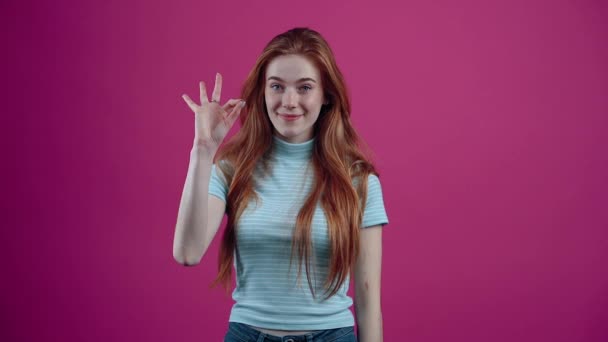 Портрет красивой, веселой рыжеволосой девушки показывает знак "хорошо" и моргает шикарными глазами. Веснушчатая девочка-подросток в голубой футболке, изолированная на розовом фоне. Концепция народов — стоковое видео