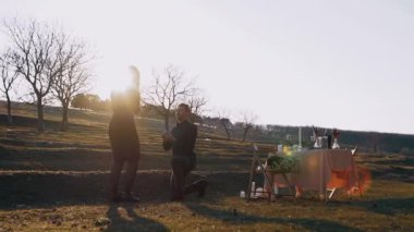 Doğada izole bir şekilde, gün batımında aşık olan adam, sevgili kadına dizlerinin üstünde evlenme teklif eder ve kadın da ellerini zevkle ağzına koyar.