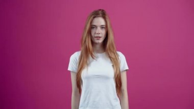 Kızıl saçlı ve karizmatik genç kadın portre işaretini gösterir, onu kaldırır, ona yaklaşır, geniş bir şekilde gülümser. Beyaz tişörtlü, pembe arka planda izole edilmiş çilli genç kız. Konsept: