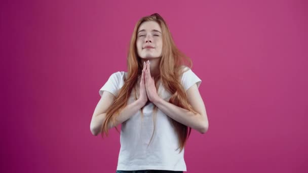 The hopeful charismatic redhead pleads her hands to her chest. Freckled teenage girl in a white T-shirt, isolated on a pink background. The concept of peoples lifestyle — Stock Video