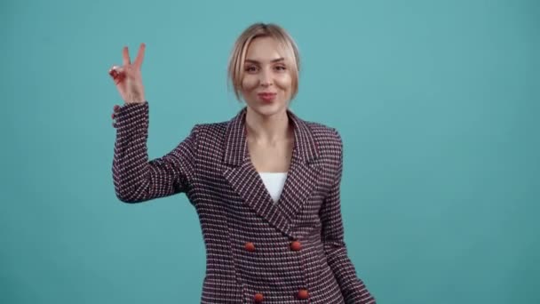 The charming, charismatic young blonde shows the signs of peace and dances energetically with them, displaying a wide smile. Isolated on a turquoise background.The concept of life. Peoples emotions — Stock Video