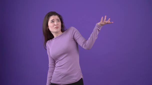 混乱した若い女性は、それが何であるかを尋ね、一方の側から他方へ彼女の手を指してから、彼女の印象的な手を与えます。紫のセーターに身を包んだ、紫の背景に隔離されたブルネット — ストック動画