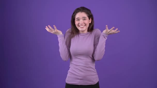 La jeune femme optimiste avec un large sourire hausse les épaules dans l'étonnement et met honteusement sa main sur son visage. Brunette isolée sur un fond violet, vêtue d'un pull violet. Concept de style de vie — Video