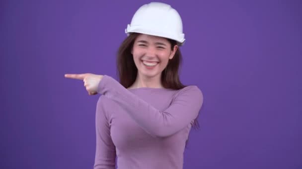 El retrato de una chica optimista en un casco blanco de ingenieros apuntando a un lado sonríe ampliamente. Morena aislada sobre un fondo morado, vestida con un suéter morado. Concepto de estilo de vida — Vídeos de Stock