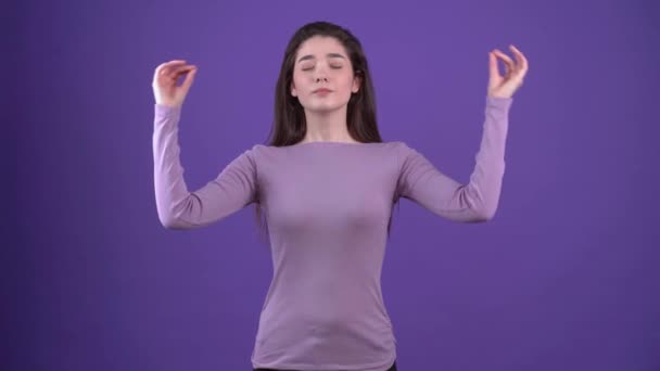 Wanita muda cantik dengan mata tertutup melakukan latihan yoga, meditasi dengan jari-jarinya bersama-sama. Dia mengenakan sweater ungu di studio dengan latar belakang ungu — Stok Video