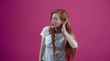 Kafası karışmış genç kadın kulağına fısıltıları duyamadığı için elini koyar. Beyaz tişörtlü, pembe arka planda izole edilmiş çilli genç kız. İnsan kavramı.