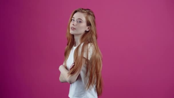 Das Porträt einer rothaarigen, charismatischen jungen Frau blickt selbstbewusst nach vorn, verschränkt die Hände auf der Brust, zeigt ein breites Lächeln. Sommersprossige Teenager-Mädchen in einem weißen T-Shirt, isoliert auf einem rosa — Stockvideo