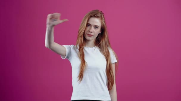 The portrait of a dissatisfied redhead who shows the sign of dislike with both hands. Freckled teenage girl in a white T-shirt, isolated on a pink background. The concept of peoples lifestyle — Stock Video
