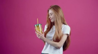 Kızıl saçlı öğrenci, elinde bir bardak meyve suyu ile tatmin olmuş, hayal kurarak gülümsüyor. Beyaz tişörtlü, pembe arka planda izole edilmiş çilli genç kız. İnsanların yaşam tarzı kavramı