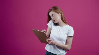 Telefon görüşmesine odaklanan genç kızıl not alır ve günlükten alınan görevleri yazar. Beyaz tişörtlü, çilli genç, pembe arka planda izole edilmiş. Konsept: