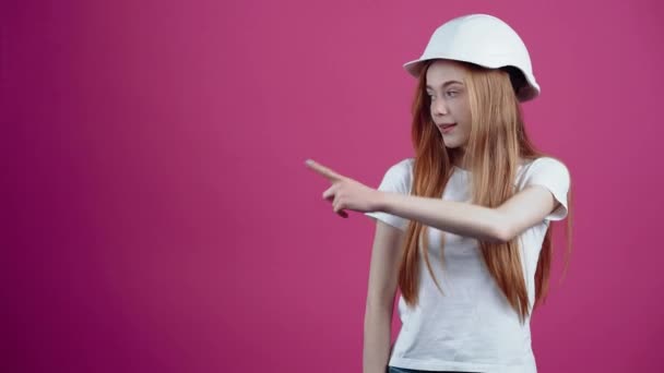 Den unge rødhåret med ingeniørhjelmen peger med pegefingeren til den ene side og godkender. Freckled teenage pige i en hvid T-shirt, isoleret på en lyserød baggrund. Begrebet folk – Stock-video