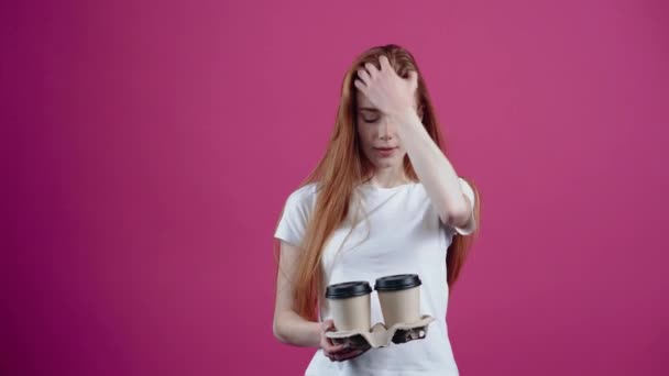 Оптимистичная и харизматичная студентка с двумя чашками кофе в руке кокетливо улыбается и протягивает их вперед, чтобы подарить кому-нибудь. Веснушчатый подросток в белой футболке, изолированный на розовом — стоковое видео