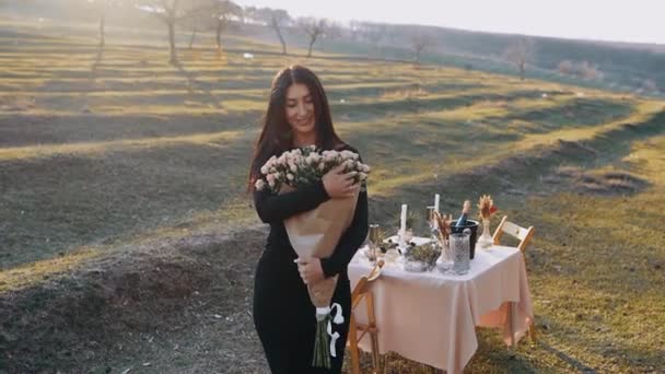 De gelukkige vrouw voelt zich geliefd met een prachtig boeket bloemen in haar armen dat ze verheven ruikt, in de boezem van de natuur met zonsondergang, tijdens een romantisch diner — Stockvideo