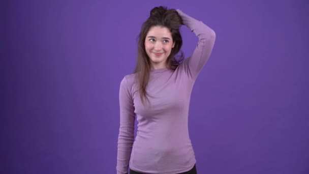 Fata carismatică, elegantă plimbându-și mâinile prin păr și privea înainte cu un leu zâmbet. Bruneta izolată pe un fundal violet, îmbrăcată într-un pulover violet. Conceptul de stil de viață — Videoclip de stoc
