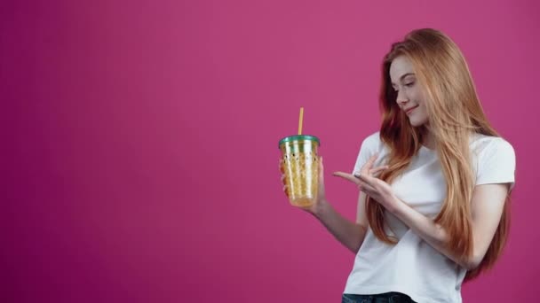 Le portrait d'une rousse charismatique avec un verre de jus à la main sourit largement et pointe du doigt le verre, et hoche la tête avec approbation. Adolescente rousse en T-shirt blanc, isolée sur un rose — Video