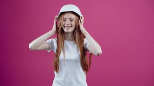 エンジニアの白いヘルメットと若い赤毛は、楽観的に笑顔と自信を持って先を見ています。ピンクの背景に隔離された白いTシャツに身を包んだ10代の少女。人々の概念 — ストック動画