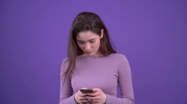 Güzel genç kadın telefonda yazı yazıyor, bir tarafa bakıyor, şokta, işaret parmağıyla işaret ediyor, sonra neşeli kadın yazmaya devam ediyor. Esmer, mor bir arka planda izole edilmiş,